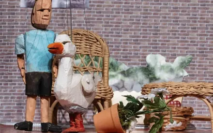 Puppentheater-Szenerie: eine Ziegelmauer im Hintergrund im Vordergrund zwei Holz-Marionetten, eine davon eine Figur mit blauem T-Shirt und schwarzen kurzen Hosen, die zweite Figur eine Gans. Im Raum noch ein Rattan-Couchtisch und ein Rattan-Sessel, davor ein umgestoßener Blumentopf mit Margeriten