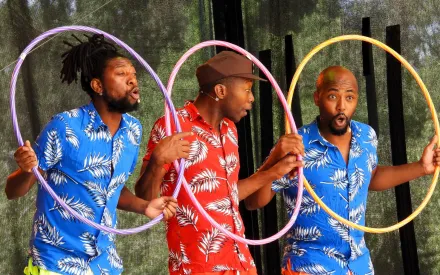 Drei Männer in Hawaiihemden mit Palmenmuster, zwei in Rot, einer in Blau, stehen nebeneinander und halten sich Hula Hoop-Reifen vor den Körper, schauen durch die Reifen