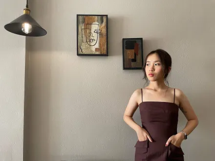 Frau mit Trägerkleid, die Hände in die Taschen gesteckt, Blick leicht zur Seite gerichtet. Die Frau steht vor einer Wand, an der 2 Bilder hängen