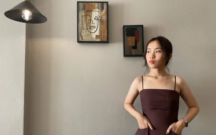 Frau mit Trägerkleid, die Hände in die Taschen gesteckt, Blick leicht zur Seite gerichtet. Die Frau steht vor einer Wand, an der 2 Bilder hängen