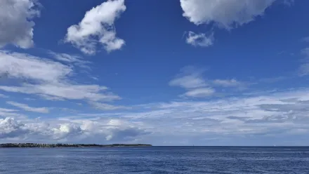Blick aufs Meer, blauer Himmel mit vereinzelten Wolken, in der Ferne am Horizont ist Land zu sehen