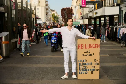 Man in weiß gekleidet, in einer Fußzone mit geschäftigem Treiben stehend, hält ein Pappkartonschild mit Aufschrift: HEILUNGEN alle Kassen! Gicht: 15€, Blindheit: 100€, Pilze: 40 €, Schmerzen: 10€, Covid: 15€