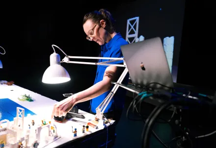Eine Frau arbeitet konzentriert an einem LEGO-Modell unter hellem Licht, neben ihr steht ein Laptop.