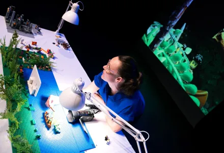 Eine Frau arbeitet an einem beleuchteten Tisch mit LEGO-Figuren und Pflanzenmodellen, während eine Projektion im Hintergrund zu sehen ist.