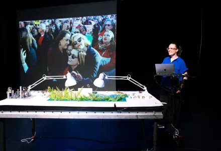 Eine Frau steht lächelnd an einem Tisch mit Miniaturlandschaft und Laptop, während im Hintergrund ein großes Foto projiziert wird.