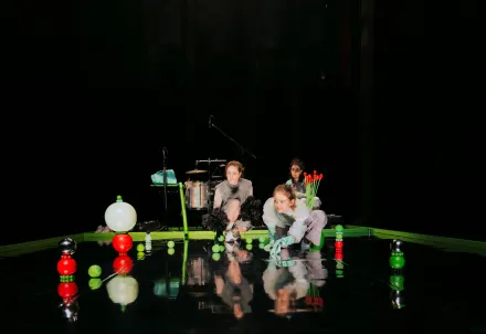 Drei Personen in hellen Kostümen agieren auf einer dunklen Bühne mit grünen und roten Kugeln; im Hintergrund stehen Schlagzeug und Mikrofone.