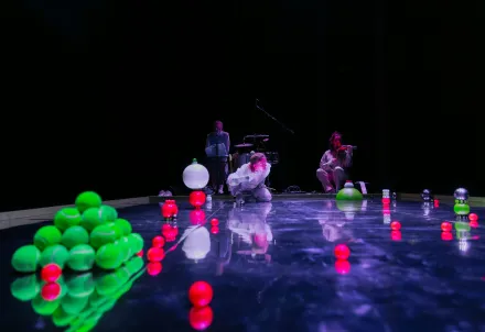Drei Personen auf einer dunklen Bühne, umgeben von leuchtend grünen und roten Kugeln; eine spielt Violine, während die anderen zwischen Instrumenten und Lichteffekten agieren.