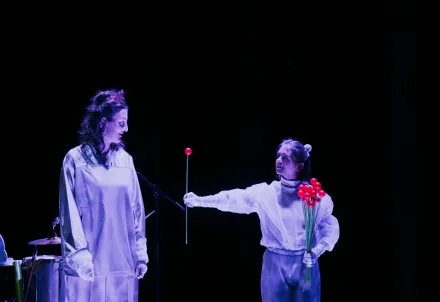 Zwei Personen in hellen Kostümen stehen auf der Bühne; eine hält einen Strauß roter Stäbe und reicht der anderen einen einzelnen roten Stab vor schwarzem Hintergrund.