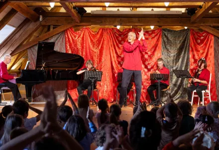 Ein Musikerensemble spielt auf einer Bühne mit rotem Vorhang vor einem applaudierenden Publikum.