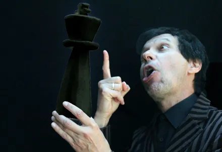 Person in gestreiftem Sakko hält eine übergroße schwarze Schachfigur in der Hand und blickt mit theatralischem Ausdruck darauf, vor schwarzem Hintergrund.