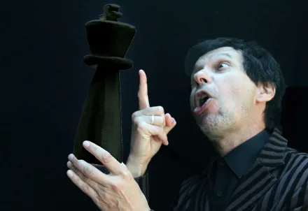 ChatGPT:  Person in gestreiftem Sakko hält eine übergroße schwarze Schachfigur in der Hand und blickt mit theatralischem Ausdruck darauf, vor schwarzem Hintergrund.