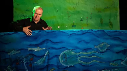 Im Hintergrund eine Leinwand, bemalt mit Wiese und Blumen, davor eine Erzählperson. Vor der Person eine Leinwand mit einer lustigen Unterwasserwelt mit unterschiedlichen Fischen
