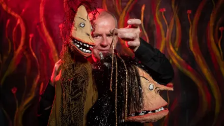 Person hält zwei ausdrucksstarke Handpuppen mit grotesken Gesichtern vor rotem, feurig wirkendem Hintergrund – eine lebendige Theaterszene voller Farbe und Kontrast.