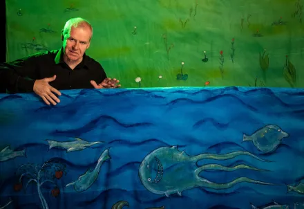 Person steht hinter einer bunt bemalten Stoffkulisse mit Meeresmotiven und Fischen im Vordergrund sowie einer grünen, pflanzenverzierten Landschaft im Hintergrund.