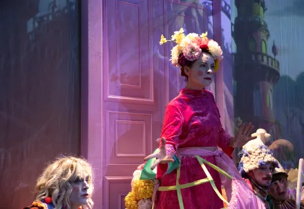 Person in pinkem Samtkleid mit großer Blumenkrone vor einer beleuchteten Tür, im Hintergrund Figuren in farbenfrohen Kostümen.