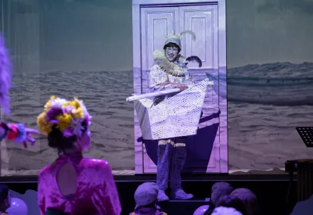 Person mit Mühlradkragen und Schiff aus beschriebenem Papier, im Hintergrund eine Produktion mit Meer, das aus Wörtern besteht