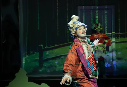 Person mit weiß geschminktem Gesicht und bunter, karierter Jacke, auf dem Kopf eine Mütze mit kleinem Schaf. Im Hintergrund eine Waldprojektion mit Zaun.