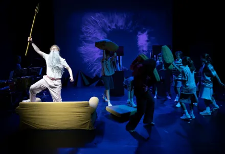 Mehrere junge Darsteller*innen toben auf der Bühne mit Kissen, während eine Figur mit weißem Bart und Dreizack auf einem Sofa steht – im Hintergrund leuchtet eine blaue Projektion wie eine Explosion oder ein Auge.