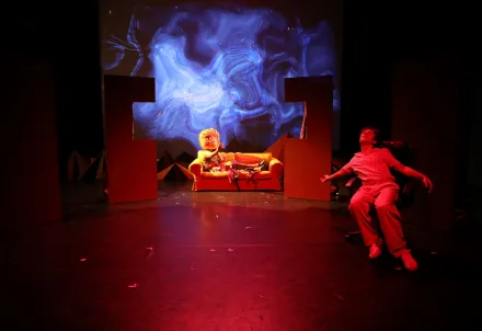 Ein Darsteller sitzt entspannt in einem Sessel, während im Hintergrund eine Figur mit übergroßem Kopf auf einem Sofa liegt; auf der Leinwand dahinter wirbeln blaue, abstrakte Formen.