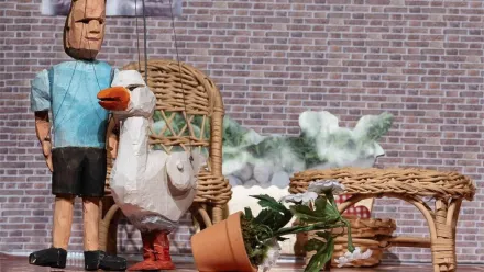 Puppentheater-Szenerie: eine Ziegelmauer im Hintergrund im Vordergrund zwei Holz-Marionetten, eine davon eine Figur mit blauem T-Shirt und schwarzen kurzen Hosen, die zweite Figur eine Gans. Im Raum noch ein Rattan-Couchtisch und ein Rattan-Sessel, davor ein umgestoßener Blumentopf mit Margeriten