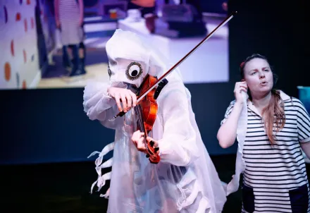 Ein fantasievoll anmutendes Unterwasserwesen spielt Geige, eine Performerin hört gebannt zu und singt mit