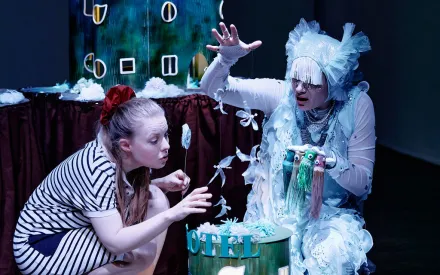 Frau in weißem, quallenartigen Kostüm kniet mit lustigen kleinen Figuren in der Hand vor einem kleinen Hotel-Modell, ein Mädchen mit schwarz-weiß gestreiftem Kleid und roten Schuhen hält kleine Windräder in der Hand und bläst in Richtung der kleinen Figuren in der Hand der Frau