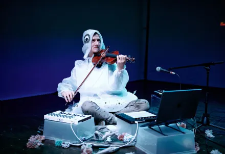 Ein Unterwasserwesen sitzt inmitten verschiedener Mischpulte und spielt Geige.