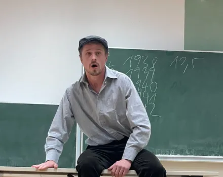 Mann sitzt auf einem Tisch vor einer Tafel mit Zahlen, trägt helles Hemd und dunkle Hose
