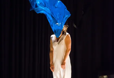 Eine Tänzerin steht auf der Bühne, während ein blauer Stoff von einem Ventilator in die Luft geweht wird.