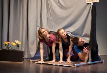 Drei Performer*innen kriechen neugierig unter einem Vorhang hervor.