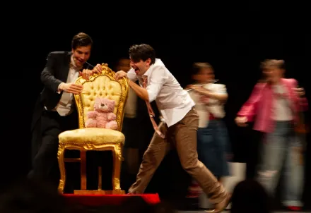 Eine dynamische Theaterszene voller Bewegung und Humor – im Mittelpunkt ein rosa Teddybär auf einem goldenen Stuhl.