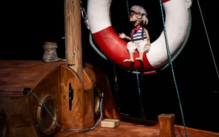 Im Mittelpunkt hängt eine Gliederpuppe mit Brille, gestreiftem Oberteil und kurzen Hosen in einem großen Rettungsring mit rotem und weißem Muster. Die Szene spielt auf einem hölzernen Schiffsdeck mit sichtbaren Bullaugen, Seilen und einem kleinen Holzkasten. Der Hintergrund ist dunkel gehalten, sodass die Figuren und das Deck deutlich hervortreten.
