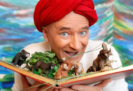 Mann mit rotem Turban hält ein Buch mit Tierfiguren und Pflanzen.