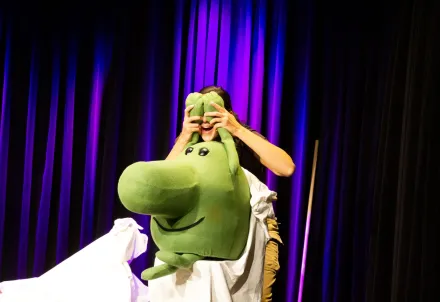 Schauspielerin hält eine große grüne Handpuppe vor sich, während sie auf der Bühne steht.
