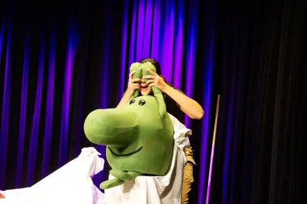 Eine lustige grüne Figur sitzt auf einem weißen Laken, dahinter steht eine Schauspielerin, lachend. Die Schauspielerin hält sich die Hände der grünen Figur vor die Augen, man sieht von ihrem Gesicht nur den lachenden Mund