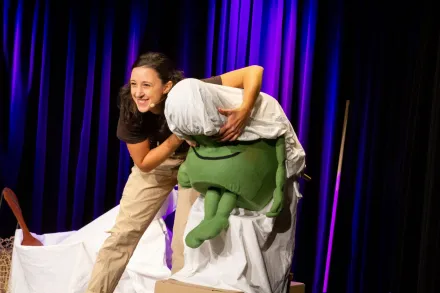 Eine Schauspielerin mit einer grünen, lustigen Figur mit großer Nase, der sie ein Laken über Augen und Nase hält.