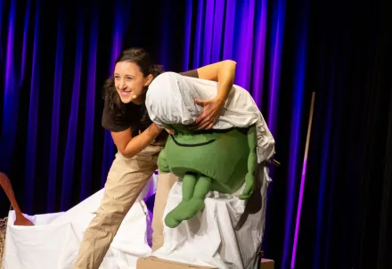 Schauspielerin lacht und bewegt eine grüne Puppe auf der Bühne vor dunklem Vorhang.