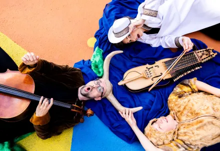 Drei Künstler:innen liegen farbenfroh auf dem Boden, umgeben von Instrumenten und Stoffen.