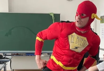 Person in rotem Superhelden-Kostüm mit gelben Blitzen und Schriftzug 'El Bueno' posiert in einem Klassenzimmer