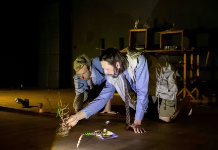 Zwei Performer knien auf dem Boden und bewegen kleine Figuren in einer beleuchteten Szene.