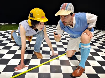 Zwei Männer knien auf einem schwarz-weiß karierten Boden. Einer trägt gelben Bauhelm, Jeans und braune Schuhe, der andere eine bunte Kappe, blaue Kniestrümpfe, weiße Shorts und braune Schuhe. Sie messen mit einem gelben Maßband.