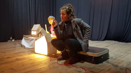 Eine Person in grauer Jacke sitzt auf einem Koffer, eine Hand in der Jackentasche, mit der anderen gestikulierend, erzählt etwas. Daneben kleine Häuser, aus deren Inneren Licht strahlt. Im Hintergrund eine zweite Person, die die erzählende Person ansieht