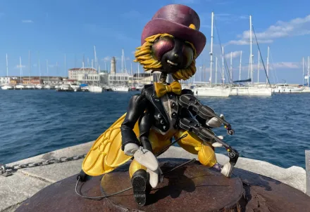 Eine detailreiche Insektenfigur mit Geige und Zylinder sitzt am Hafen vor Segelbooten.