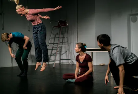 Vier Darsteller:innen interagieren lebhaft auf der Bühne, eine Person springt in die Luft.