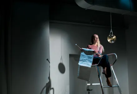 Eine Schauspielerin sitzt auf einer Leiter, hält eine Papierrolle und blickt nachdenklich in den Raum.